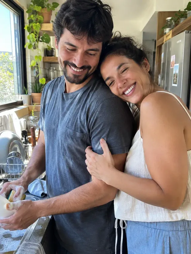 Marido lavando a louça e esposa sorrindo abraçada a ele
